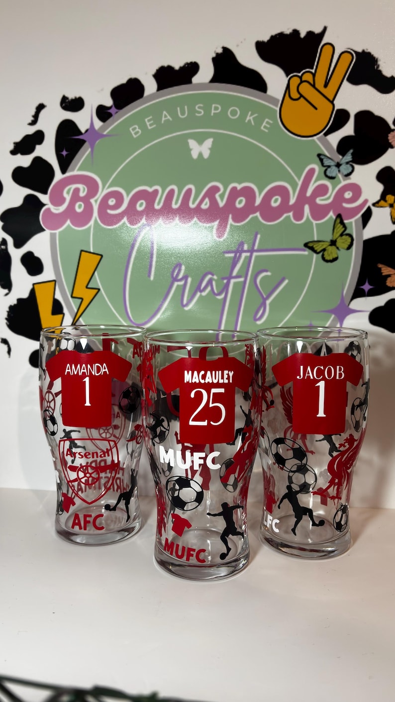 May include: Three clear pint glasses with red soccer jersey designs. Each glass features a name and number, with team logos and soccer ball graphics. The names are Amanda, Macauley, and Jacob. The background includes a logo that says "Beauspoke Crafts".