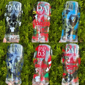 May include: Six clear pint glasses with personalized designs. Each glass features a name, sports-themed graphics, and the word "DAD" or a number. The designs include soccer balls, jerseys, and a lion. The glasses are displayed against a green background.