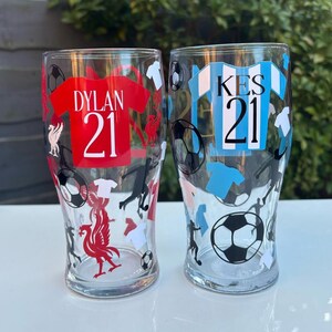 May include: Two clear pint glasses with soccer-themed designs. One glass features a red jersey with "DYLAN 21", the other a blue jersey with "KES 21". Both glasses have soccer balls, player silhouettes, and team logos.