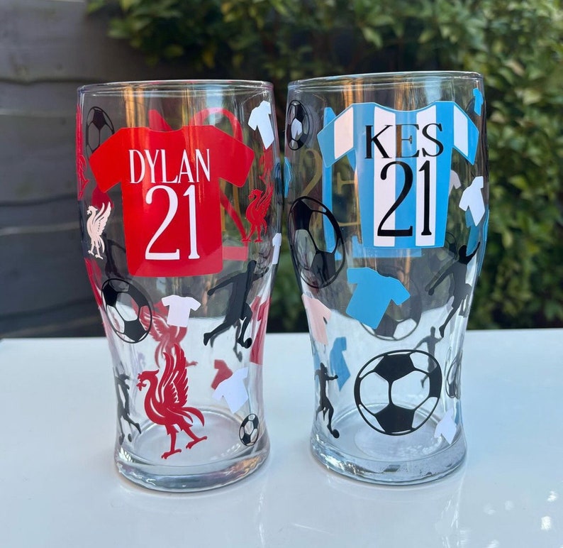 May include: Two clear pint glasses with soccer-themed designs. One glass features a red jersey with "DYLAN 21", the other a blue jersey with "KES 21". Both glasses have soccer balls, player silhouettes, and team logos.