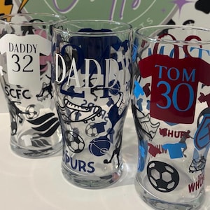 May include: Three clear pint glasses with soccer-themed designs. One glass has "DADDY 32" on a white jersey, another has "DADDY" in navy blue, and the third has "TOM 30" on a maroon jersey. Each glass features soccer ball and shoe graphics.
