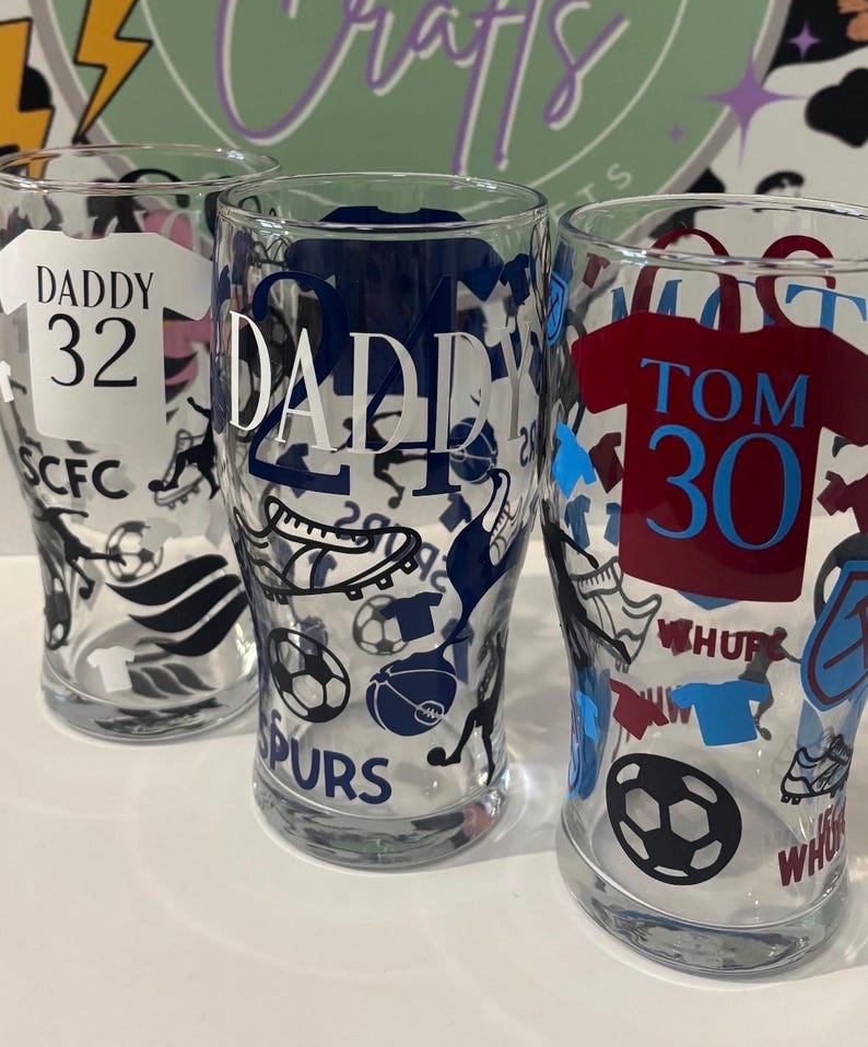 May include: Three clear pint glasses with soccer-themed designs. One glass has "DADDY 32" on a white jersey, another has "DADDY" in navy blue, and the third has "TOM 30" on a maroon jersey. Each glass features soccer ball and shoe graphics.
