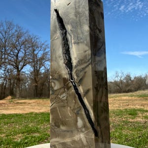 May include: A tall, rectangular stone obelisk with a pointed top. The stone is primarily beige and gray with dark gray veining and a prominent black fissure running down the front. The obelisk is outdoors, set against a backdrop of a blue sky and green grass.