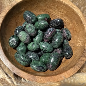 May include: A wooden bowl filled with polished zoisite stones. The stones are oval-shaped, with a mix of dark green, black, and pinkish-red hues. The bowl is made of light-colored wood, and the background is blurred.