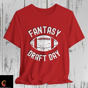 May include: A red short-sleeved t-shirt featuring a white graphic of an American football in the center. The text "FANTASY" is printed in white above the football, and "DRAFT DAY" is printed in white below it, both in a distressed font.