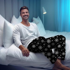 May include: Black pajama pants with a white snowflake pattern. The pants are part of a sleepwear set. The image shows the pants on a person sitting on a bed. The person is wearing a white long-sleeve shirt.