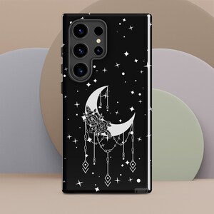 Puede incluir: Carcasa de teléfono negra con un diseño de luna creciente blanca con detalles florales y una cadena que cuelga de ella. El fondo es una noche estrellada negra.