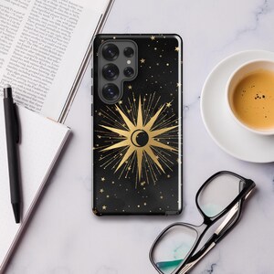 Puede incluir: Una funda de teléfono negra con un diseño de sol y luna dorados y una dispersión de estrellas doradas. La funda del teléfono tiene un recorte para el objetivo de la cámara.