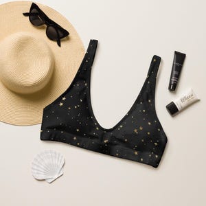 May include: Black bikini top with a gold star pattern. The top is a scoop neck style. A straw hat, sunglasses, sunscreen, and a seashell are also in the image.