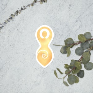 May include: Gold sticker with a stylized female figure with a spiral inside. The figure is in a standing pose with arms raised above the head.