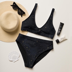 May include: Black bikini set with a celestial pattern of gold moons and stars. The set includes a top and high-waisted bottoms. Accessories include a straw sun hat, sunglasses, sunscreen, and a seashell.