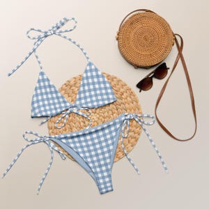 May include: A blue and white gingham bikini set with tie fastenings. The swimwear is displayed with a round woven bag, sunglasses, and a woven placemat. The bikini top and bottom are made of a soft, lightweight fabric.