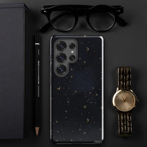 Puede incluir: Una funda de teléfono negra con un fondo azul oscuro y un patrón de estrellas doradas y lunas crecientes. La funda tiene un recorte para la cámara con tres lentes.