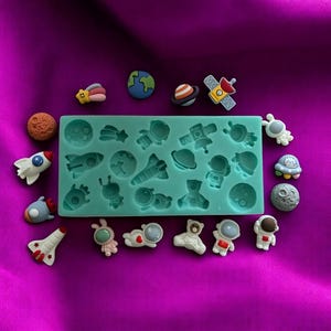 May include: A silicone mold with 16 different space-themed designs, including astronauts, rockets, planets, and stars. There are also several finished resin pieces in the same designs, including a blue and green planet, a red and white rocket, and a white astronaut with a red heart.