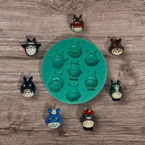 Peut inclure: Un moule en silicone avec neuf cavités en forme de personnage de dessin animé. Le moule est vert et les cavités sont remplies de petites figurines colorées du même personnage. Les figurines sont en résine et ont des couleurs et des détails différents.