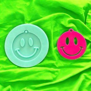 May include: Two smiley face shaped silicone molds, one is light blue and the other is pink. The molds are on a bright green surface.