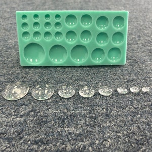 May include: A green silicone mold with 20 round cavities of varying sizes. The mold is used to create resin cabochons. There are 10 clear resin cabochons of varying sizes in a row on a gray surface.