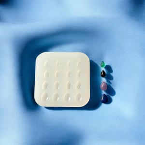 May include: A white silicone mold with 16 teardrop-shaped cavities.  Next to the mold are four teardrop-shaped gemstones in green, black, purple, and blue.
