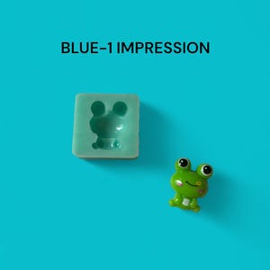 May include: A green frog figurine with a white smile and black eyes. The frog is sitting on a blue background. The image also shows a silicone mould for making a frog figurine. The mould is light blue and has a frog shape.