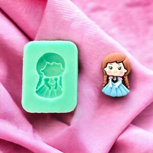 May include: A green silicone mold with a cutout of a cartoon girl with brown hair wearing a blue dress. Next to the mold is a small, blue and brown figurine of the same cartoon girl.