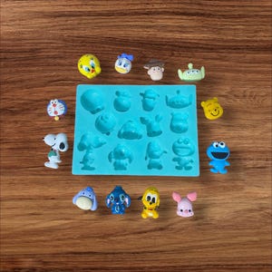 May include: A blue silicone mould with 16 different cartoon character shapes. The mould is surrounded by small figurines of the characters, including Tweety Bird, Donald Duck, Woody, Buzz Lightyear, Winnie the Pooh, Snoopy, Stitch, and more.