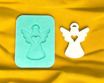May include: A white silicone mold for making angel ornaments. The mold is shaped like an angel with wings and a heart in the center. The mold is on a yellow surface.
