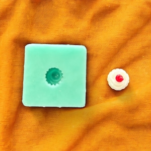 May include: A green silicone mould for making a small cake with a single red cherry on top. The mould is on a yellow fabric background.