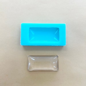 Puede incluir: Un molde de silicona azul claro con una cavidad rectangular para hacer manualidades de resina. Una pieza rectangular de resina transparente se muestra junto al molde.