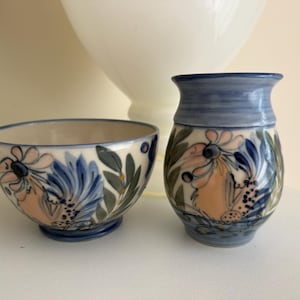 May include: A ceramic bowl and a matching small vase featuring a hand-painted floral pattern in shades of blue, soft peach, and sage green on a cream-coloured background. Both pieces have a smooth, glazed finish and a flared rim design.