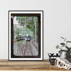 Pode incluir: Fotografia emoldurada de um trem se aproximando de uma estação. O trem é branco e azul, viajando em trilhos por uma paisagem verde. A imagem é emoldurada com uma borda preta.