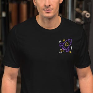 May include: Black t-shirt featuring a purple rocket ship graphic with a yellow flame and stars. The rocket ship has a heart shape inside. The shirt is made of a soft material.