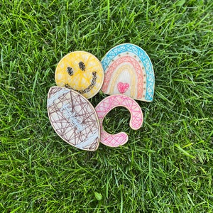 May include: Four wooden string art pieces on a green grassy background. The pieces are a smiley face, a rainbow with a heart, a football, and a letter C. The smiley face is yellow with black eyes and a mouth. The rainbow is blue, pink, orange, and yellow with a red heart in the center. The football is brown and white. The letter C is pink.