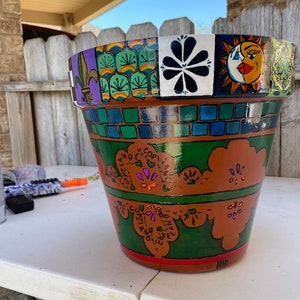 Puede incluir: Una maceta de terracota pintada con diseños coloridos, incluyendo un sol y una luna, una flor de lis y un patrón floral. La maceta está decorada con pintura verde, naranja, morada y blanca.