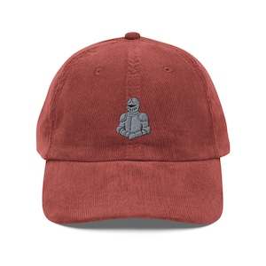 May include: A brick-red corduroy baseball cap with a curved brim. The cap features a small, embroidered image of a knight in gray armor on the front. The hat has a classic, unstructured design.