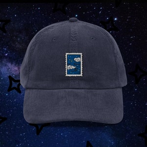 Puede incluir: Una gorra de béisbol de pana azul marino con un parche bordado blanco que representa un cielo nocturno con nubes y estrellas. El parche tiene la forma de un sello de correos.