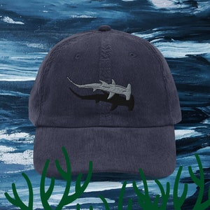Puede incluir: Una gorra de béisbol de pana azul marino con una imagen bordada de un tiburón martillo. El tiburón es gris y negro y nada en una ola de mar estilizada.
