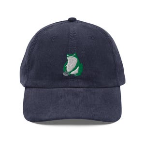 May include: Navy blue corduroy baseball cap featuring an embroidered green frog design. The frog has a white belly and a grumpy expression. The cap has a curved brim and an adjustable strap at the back.