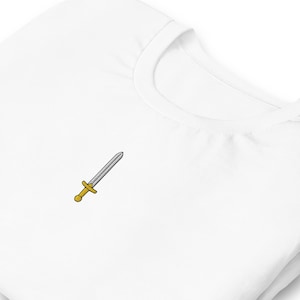 以下が含まれることがあります： 小さな剣の刺繍が施された白いTシャツ。剣は銀色の刃と金色の柄をしています。Tシャツは柔らかく快適な素材で作られています。