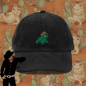 Könnte beinhalten: Eine schwarze Baseballkappe aus Cord mit einer gestickten grünen Frosch, der einen braunen Cowboyhut trägt. Der Frosch schaut nach vorne und hat den Mund geschlossen. Die Kappe befindet sich vor einem Hintergrund mit einem sich wiederholenden Muster von Fröschen, die Cowboyhüte tragen.