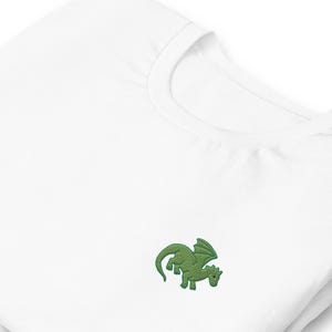 Peut inclure: Un t-shirt blanc à col rond avec un petit dragon vert brodé sur la poitrine. Le dragon a des ailes et une longue queue. Le t-shirt est fait d'une matière douce et confortable.