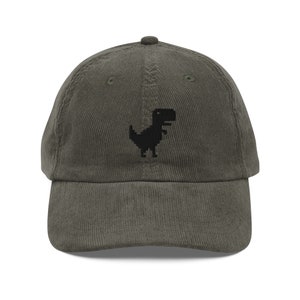 Pode incluir: Um boné de beisebol de veludo cotelê verde escuro com um dinossauro pixelado bordado em preto na frente.