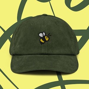 Peut inclure: Casquette de baseball en velours côtelé vert foncé avec une visière incurvée. La casquette présente une abeille de dessin animé brodée en noir, jaune et blanc sur le devant. Le fond est jaune clair avec des formes abstraites vertes.