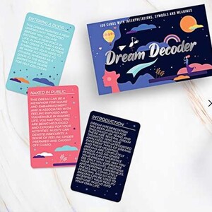 Puede incluir: Un mazo de cartas "Dream Decoder" con interpretaciones, símbolos y significados. La caja es azul marino con el título en escritura blanca. Se ven tres cartas, incluyendo una que dice "Entering a Door" y otra que dice "Naked in Public."