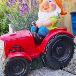 May include: A whimsical garden gnome figurine driving a bright red tractor. The gnome wears a white beard, orange hat, and green shirt. The tractor has black tires and is set against a backdrop of purple flowers and a wooden surface.