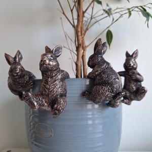 Könnte beinhalten: Vier bronzefarbene Hasenfiguren, die am Rand eines hellblauen Keramiktopfes sitzen. Die Hasen haben detaillierte Fellstrukturen und umgeben einen kleinen Baum. Der Topf hat eine strukturierte Oberfläche und ein kleines, rundes Emblem.