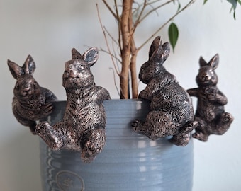 Peut inclure: Quatre figurines de lapins couleur bronze accrochées au bord d'un pot en céramique bleu clair. Les lapins ont des textures de fourrure détaillées et entourent un petit arbre. Le pot a une surface texturée et un petit emblème circulaire.