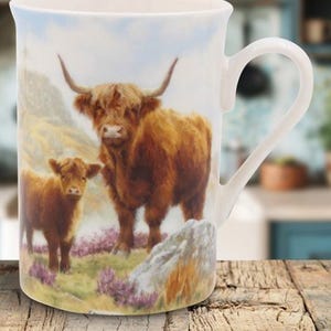 May include: White ceramic mug with a detailed painting of a Highland cow and calf in a mountainous landscape. The artwork depicts the animals' brown fur and long horns, set against a backdrop of green grass, purple flowers, and a blue sky.