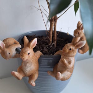 Könnte beinhalten: Ein grauer Keramik-Pflanztopf mit einem kleinen Baum und vier braunen Hasenfiguren, die an der Seite hängen. Die Hasen haben rosa Ohren und sind in verschiedenen Posen, was der Pflanze einen skurrilen Touch verleiht.