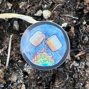 May include: A round, black and blue sticker with a watercolor illustration of two marshmallows on sticks over a campfire.