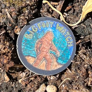 May include: A round sticker with the text "BIG FOOT WATCH" encircling an image of Bigfoot walking in a forest scene. The sticker has a black border and is set against a backdrop of soil and leaves.
